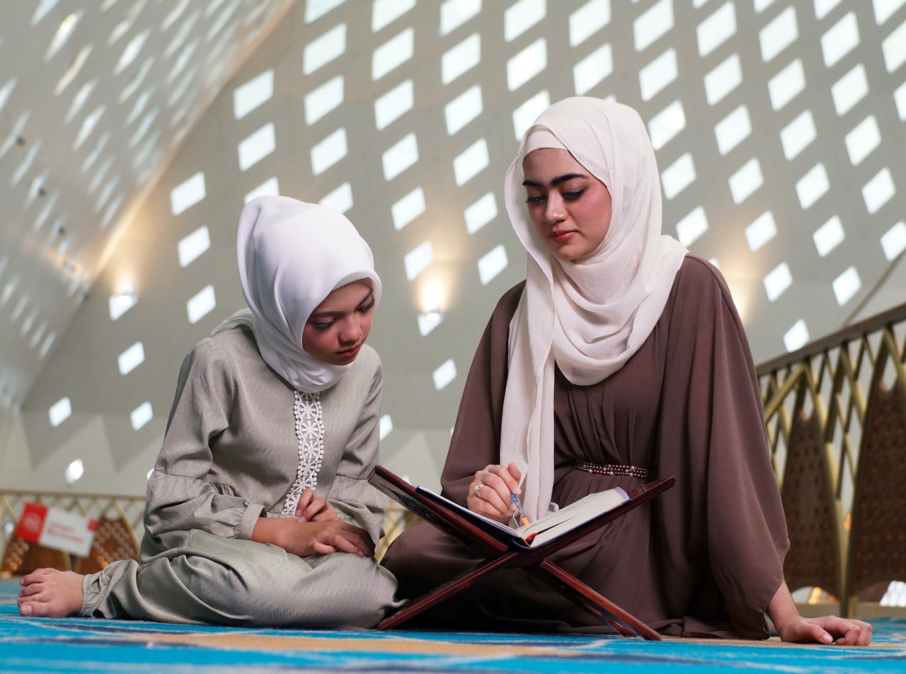 A mother and daughter in hijabs reading together inside a beautifully lit mosque.