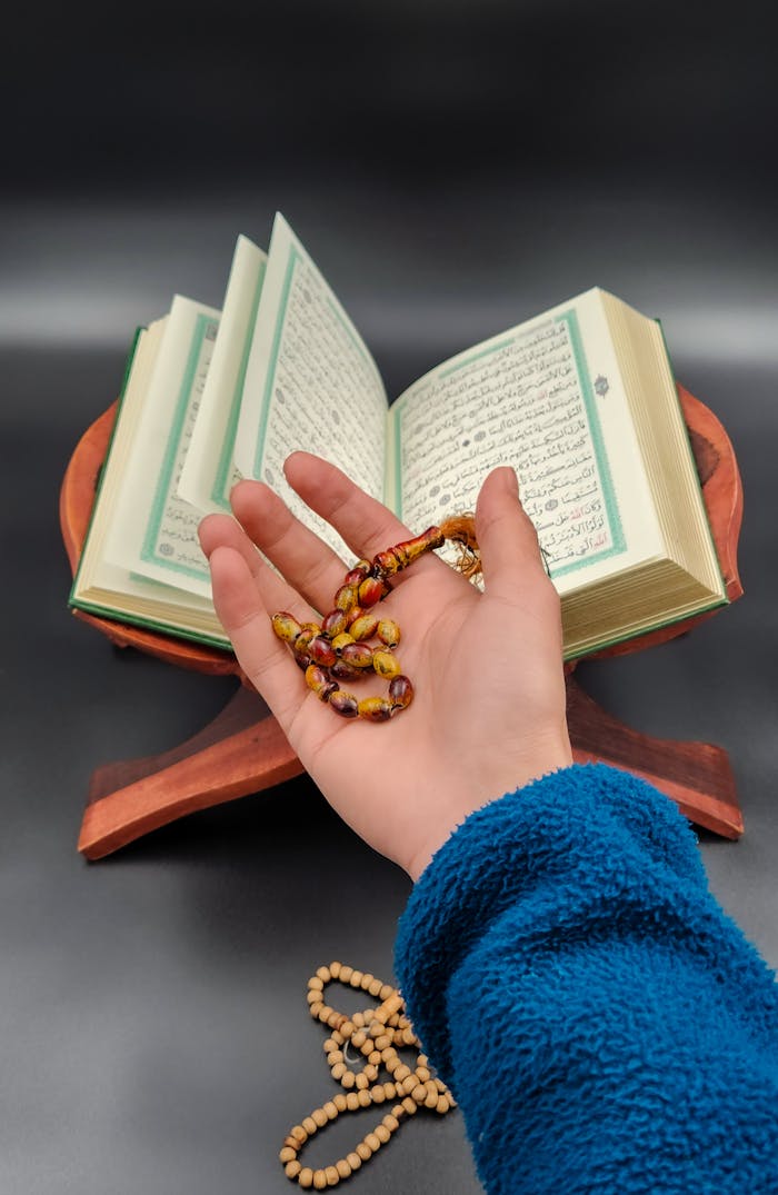 A hand holding prayer beads beside an open Quran, symbolizing spirituality and devotion.