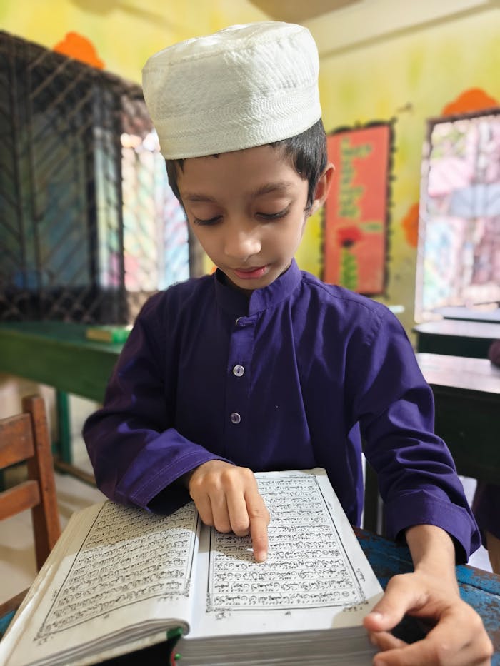 Young boy in traditional attire reading a religious book in a classroom setting.